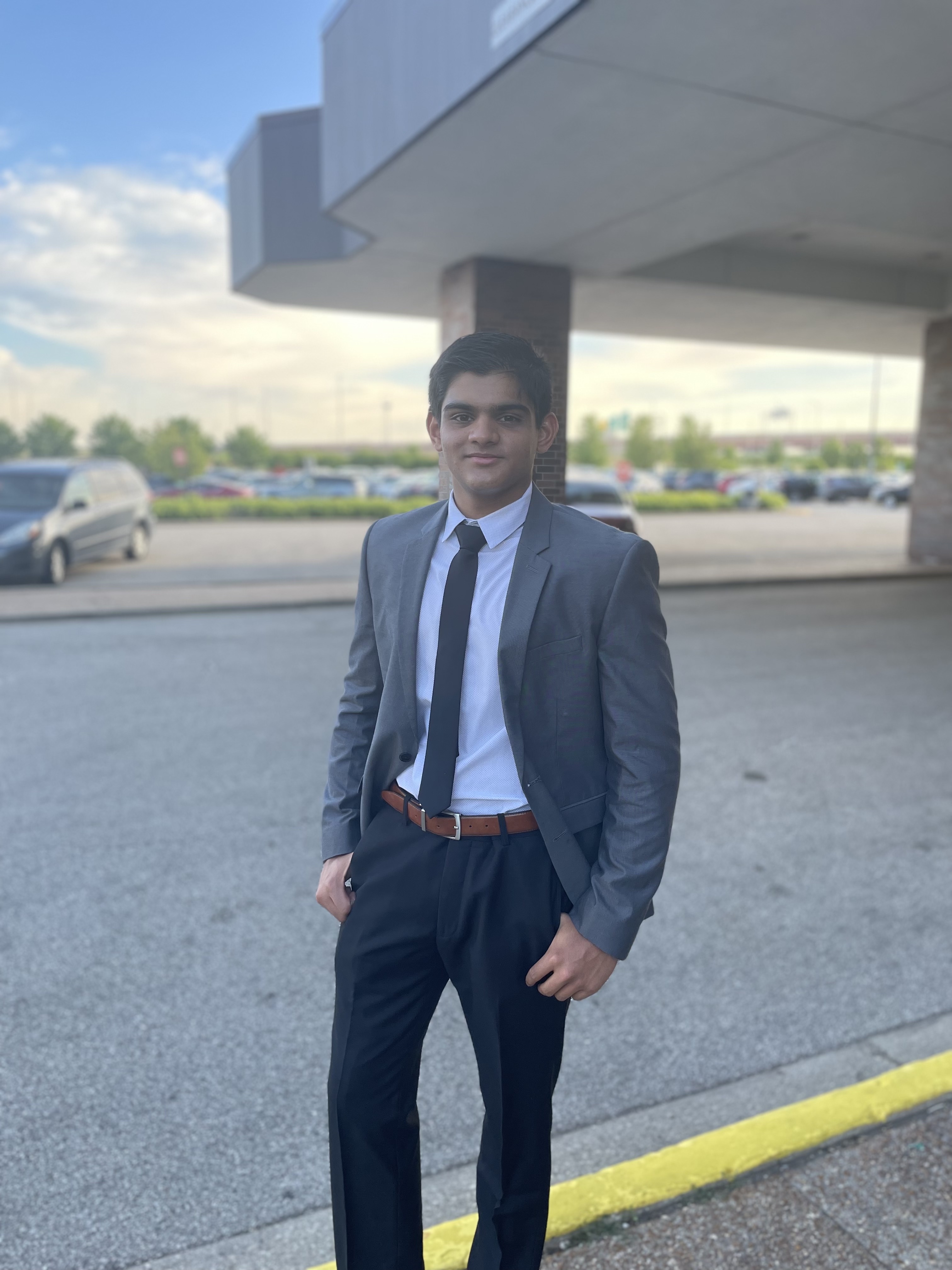Siddhant Desai Person in a formal gray suit with a tie stands smiling under a building overhang, with a parking lot and cars visible in the background.